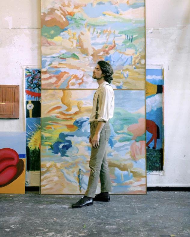 Cette photographie est l’oeuvre du photographe Raul Guillermo. Cette photographie représente le portrait en pied d’un artiste, prise dans son atelier, car il pose devant des toiles peintes. Le model est de profil, il lève légèrement la tête. Le photographe est le photographe de reportage et de mode appelé Raul Guillermo. Raul Guillermo est un photographe qui est dans l’équipe des artistes de l’agence ANTIGNY AGENCY. Cette agence d’artistes, ANTIGNY AGENCY a été créée par Alexis Antigny.