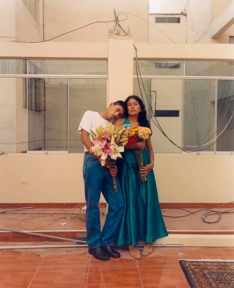 La photographie représente une mère et son fils. Les deux ont des bouquets de fleurs dans les mains. Le fils pose sa tête sur l’épaule de sa mère qui porte une robe bleue. Ils sont devant un immeuble.