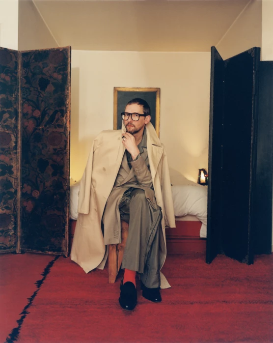 Cette photographie est le portrait du fashion editor Samuel François. Ce portrait réalisé pour le magazine ODDA a été réalisé par le photographe Raul Guillermo. Le model est assis sur une chaise, il porte un long imper. Il porte aussi des chausettes rouges qui rappellent le sol feutré de rouge lui aussi.