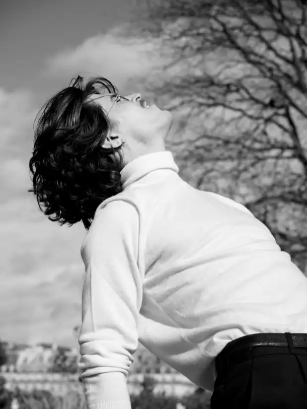 Ce visuel est une photographie en noir et blanc. La photographie est prise en contre plongé. Cette photographie est un portrait d’un mannequin en mouvement. Le mannequin est de profil et se penche en arrière comme pour évoquer une danse. La photographie a été prise aux jardins des tuilleries à Paris, France par le photographe de mode Raul Guillermo.