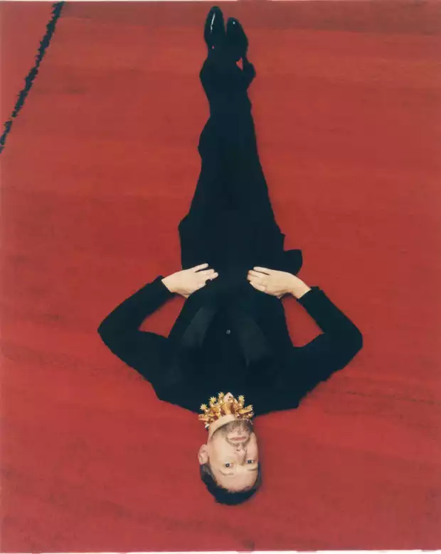 Cette photographie est le portrait du fashion editor spécialisé en bijoux appelé Samuel François.  Le modèle est photographié en top shot, il est allongé à même le sol sur un grand tapis rouge, la tête vers le bas. il porte un costume noir et un imposant collier en or. Ce portrait a été réalisé par le photographe de mode et de reportage Raul Guillermo. Le photographe Raul Guillermo a réalisé ce visuel pour le magazine ODDA. Raul Guillermo est un photographe représenté par l’agence ANTIGNY AGENCY. ANTIGNY AGENCY est une agence qui représente des artistes, notamment des photographes et des set designers. Le créateur de l’agence ANTIGNY AGENCY est un ancien acheteur d’art appelé ALEXIS ANTIGNY