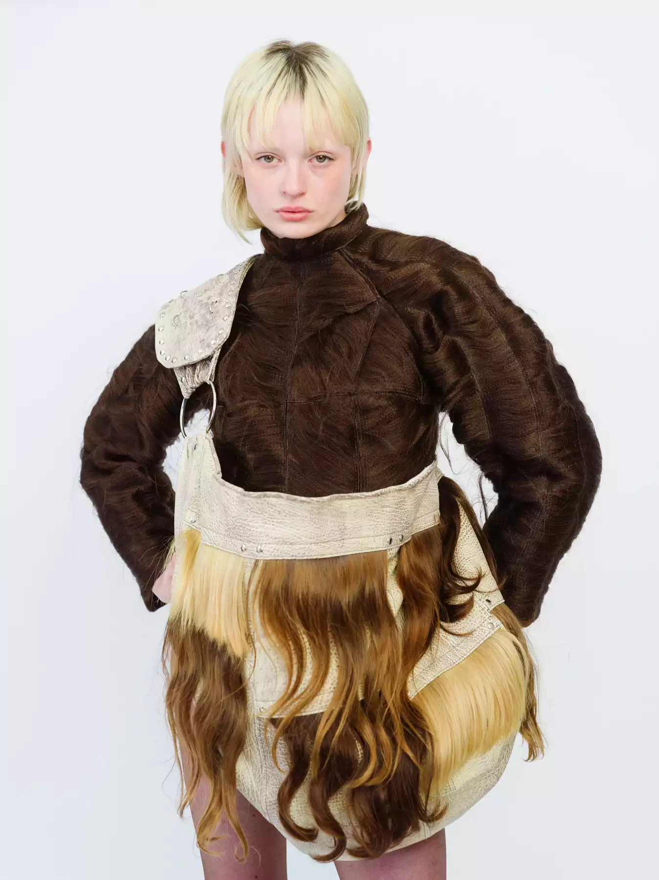 Cette photographie représente un portrait en buste d’une mannequin sur un fond blanc, en studio. La mannequin est habillée d’une tenue de couleurs marrons. Elle porte un sac étonnant où l’on semble voir des mêches de cheveux sur la bandoulière. Cette photographie a été réalisée par le photographe de mode Raul Guillermo. Raul Guillermo est un photographe de mode d’origine péruvienne représenté par l’agence d’artistes ANTIGNY AGENCY. ANTIGNY AGENCY est une agence qui représente des photographes et des set designers. Son fondateur est un ancien acheteur d’art appelé Alexis Antigny.