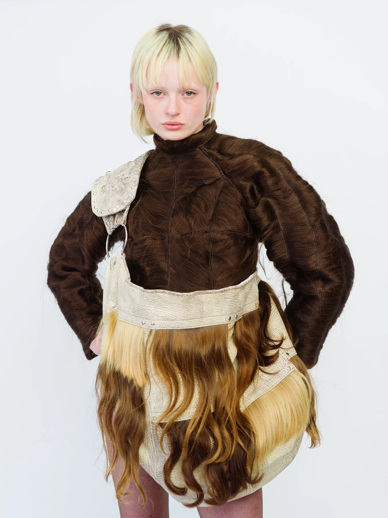 Cette photographie représente un portrait en buste d’une mannequin sur un fond blanc, en studio. La mannequin est habillée d’une tenue de couleurs marrons. Elle porte un sac étonnant où l’on semble voir des mêches de cheveux sur la bandoulière. Cette photographie a été réalisée par le photographe de mode Raul Guillermo. Raul Guillermo est un photographe de mode d’origine péruvienne représenté par l’agence d’artistes ANTIGNY AGENCY. ANTIGNY AGENCY est une agence qui représente des photographes et des set designers. Son fondateur est un ancien acheteur d’art appelé Alexis Antigny.