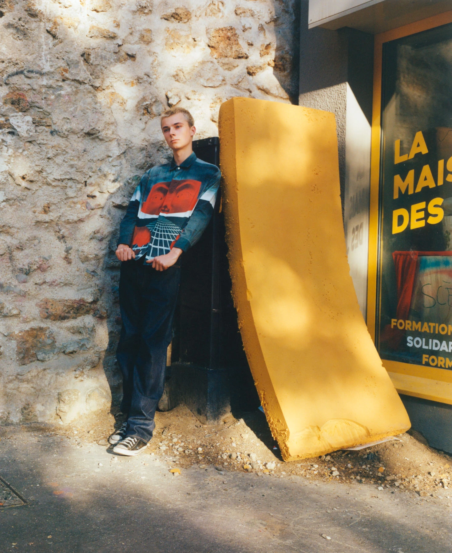 Cette photographie représente le portrait d’un mannequin blond en pied. Le mannequin porte une chemise bleu et rouge, il est adossé à un matelas jaune. Ainsi il y a tout un jeu photographique de contrastes des couleurs primaires. Le soleil vient illuminer l’ensemble de la scène. Cette photographie a été photographiée en extérieur et avec la technique photographique de l’argentique. L’auteur de cette photographie est le photographe de mode Raul Guillermo. Raul Guilllermo fait parti des photographes et artistes que représente ANTIGNY AGENCY.
