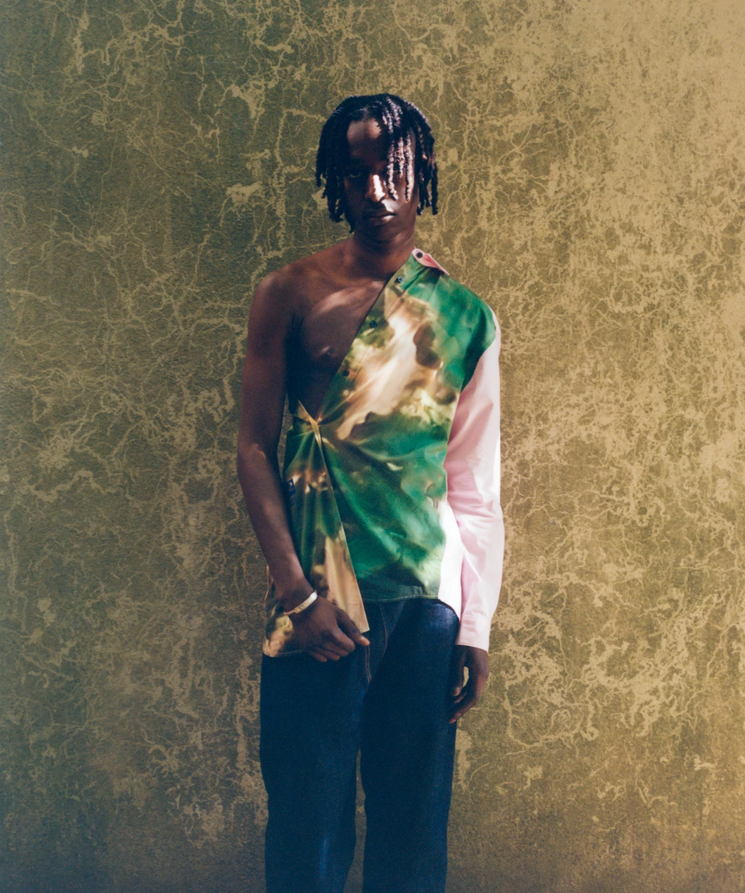 Cette photographie représente un jeune mannequin photographié contre un mur au couleurs vertes et ocres. Le mannequin porte une chemise, ou plutôt plusieurs chemises dans les tons de vert, de beige et de rose. La lumière du soleil vient éclairer le mur et surtout le visage du mannequin. Cette photographie est l’oeuvre du photographe de mode Raul Guillermo. Ce photographe de mode est d’origine péruvienne. Il fait parti de l’équipe des artistes de l’agence ANTIGNY AGENCY.